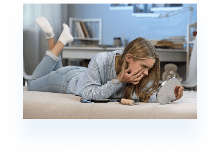 Young woman laying on her bed and touching her face while looking at herself in a small handheld mirror